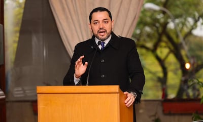 Luis Bello gesticula en podio con abrigo negro y corbata morada, frente a un ambiente formal con vegetación de fondo.