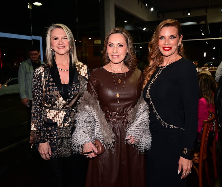 Tres mujeres sonrientes: una en chaqueta estampada, otra en vestido marrón y la tercera en vestido negro con accesorios brillantes.