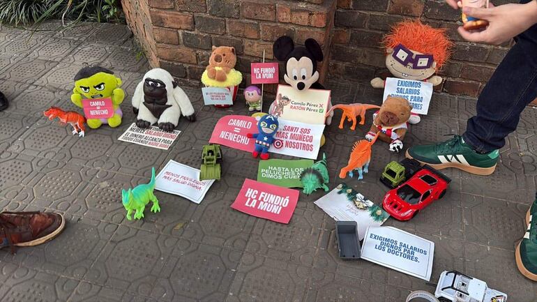Peluches de Hulk y Mickey Mouse sobre baldosas, con mensajes de protesta visibles y una mano adulta en la escena.