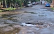 Calle deteriorada en Encarnación con baches y charcos de agua, rodeada de vehículos y vegetación.