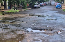 Calle deteriorada en Encarnación con baches y charcos de agua, rodeada de vehículos y vegetación.