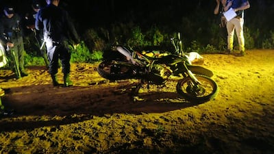 Cuatro agentes de policía en un camino de tierra, uno revisa el suelo mientras otro sostiene un arma. Motocicleta caída en el entorno.