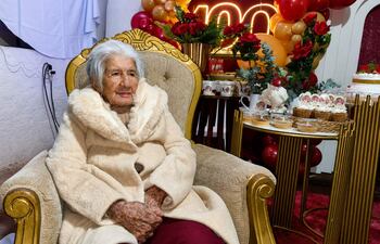 Mujer anciana con abrigo claro y cabello canoso, sentada en una silla dorada, rodeada de decoraciones festivas.