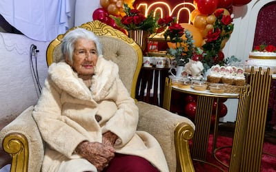 Mujer anciana con abrigo claro y cabello canoso, sentada en una silla dorada, rodeada de decoraciones festivas.