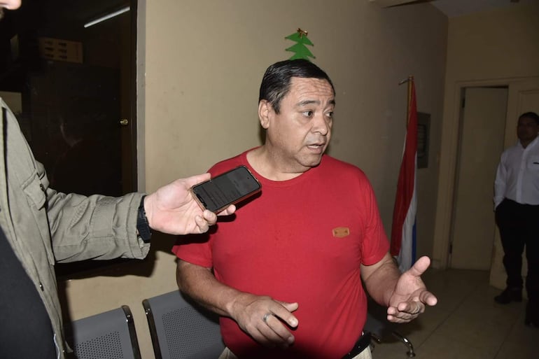 Hombre de camiseta roja y pantalones claros sostiene un teléfono, mientras un segundo hombre lo entrevista en un ambiente decorado.