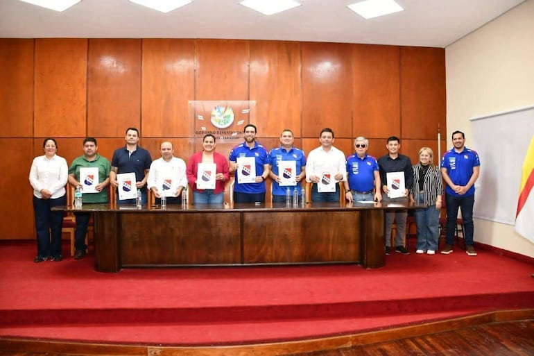 Diez personas de pie, en camisetas azules, sosteniendo documentos en una reunión formal con fondo de madera.