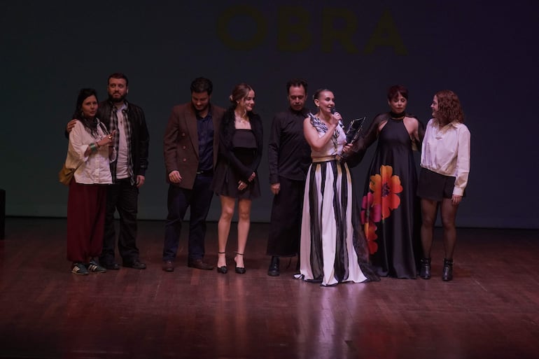 Mujer en vestido blanco y negro habla en el escenario, rodeada de artistas con diversos atuendos y flores en el auditorio.