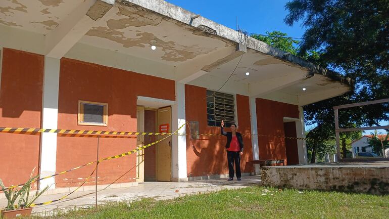 Persona con abrigo negro y camiseta roja señala la entrada de la escuela clausurada, rodeada de cintas de seguridad.
