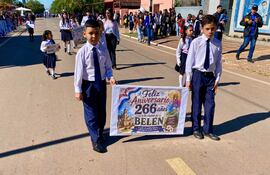 Cuatro niños en uniformes escolares sostienen un cartel con el mensaje de aniversario, sonriendo en un ambiente festivo con adultos al fondo.