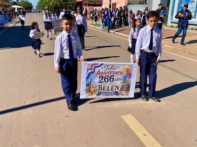 Cuatro niños en uniformes escolares sostienen un cartel con el mensaje de aniversario, sonriendo en un ambiente festivo con adultos al fondo.