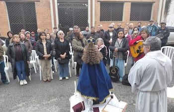 Grupo de jubilados con abrigo asiste a misa al aire libre frente a un altar sencillo, un hombre en hábito religioso dirige el acto.