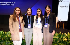 Cuatro mujeres sonrientes y vestidas formalmente posan juntas en un evento profesional con fondo decorativo y logos corporativos.