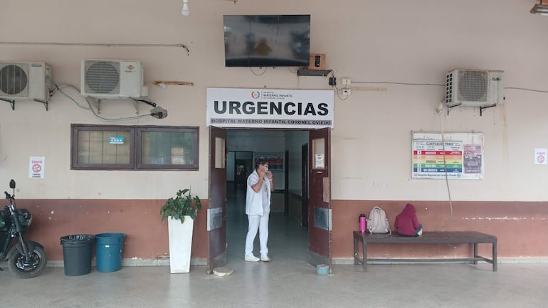 Personal médico en uniforme blanco en la entrada del hospital, mientras una persona con abrigo rojo está sentada en un banco.
