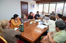 Grupo de ocho personas en sala de reuniones, escuchando atentamente a un hombre en camisa negra, con documentos y tetera sobre mesa ovalada.