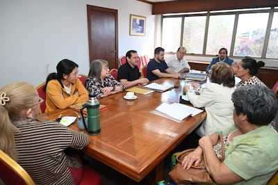 Grupo de ocho personas en sala de reuniones, escuchando atentamente a un hombre en camisa negra, con documentos y tetera sobre mesa ovalada.