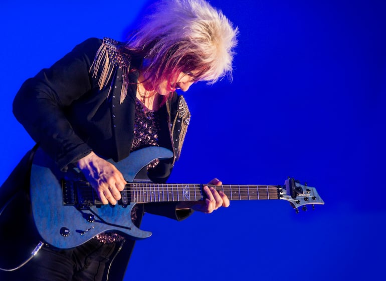 Jennifer Batten con cabello rubio y mechas coloradas, vestida de oscuro, tocando guitarra eléctrica en un escenario iluminado en azul.