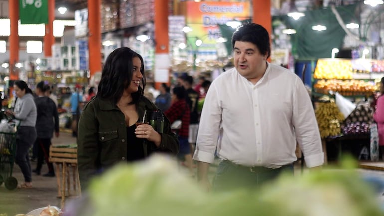 Ana Lezcano con camisa negra sosteniéndose una bebida y Jorge Montefilpo en camisa blanca, conversando en un concurrido mercado interior.