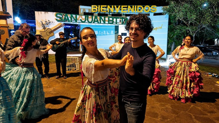 Bailarina en el centro con vestido colorido, rodeada de otros bailarines en un ambiente festivo bajo luces nocturnas.