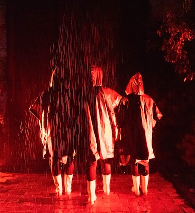 Tres figuras con ponchos oscuros se giran de espaldas, iluminadas con luces rojas en un ambiente dramático y húmedo.