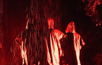 Tres figuras con ponchos oscuros se giran de espaldas, iluminadas con luces rojas en un ambiente dramático y húmedo.