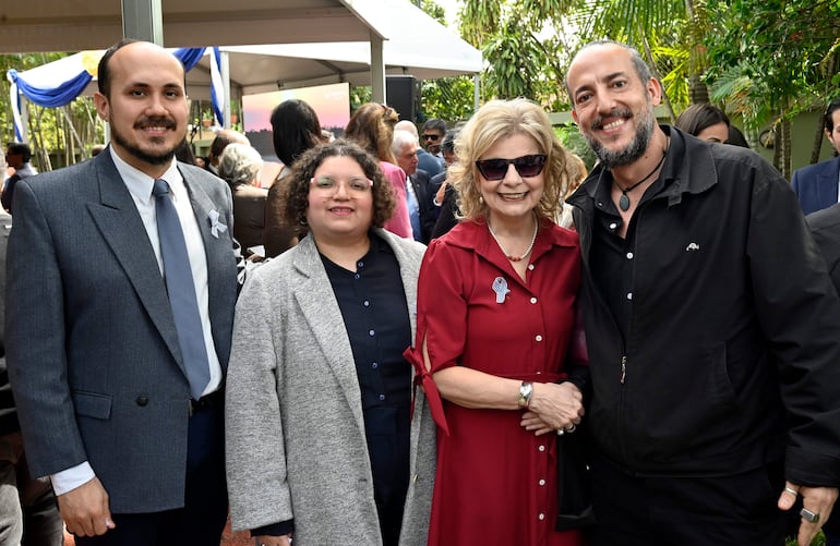 Grupo de cuatro personas sonriendo en un evento al aire libre, con un ambiente verde y festivo. Hombre con traje gris y mujer con abrigo claro.