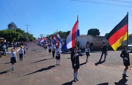 Un grupo de escolares en uniforme blanco y pantalones cortos lleva banderas, observados por adultos en un ambiente festivo y despejado.