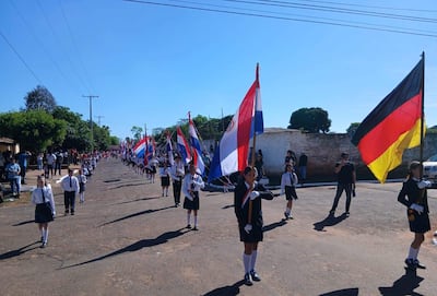 Un grupo de escolares en uniforme blanco y pantalones cortos lleva banderas, observados por adultos en un ambiente festivo y despejado.