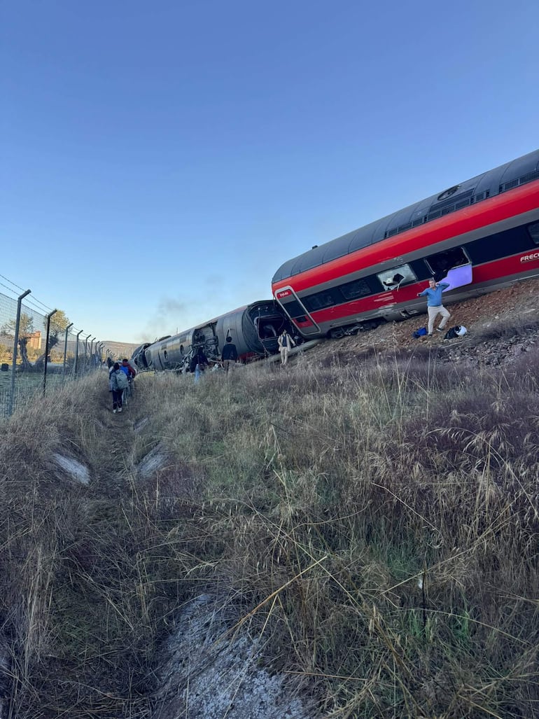 Personas intentando ayudar tras el accidente de tren, con un tren volcado de fondo y humo saliendo de un vagón.