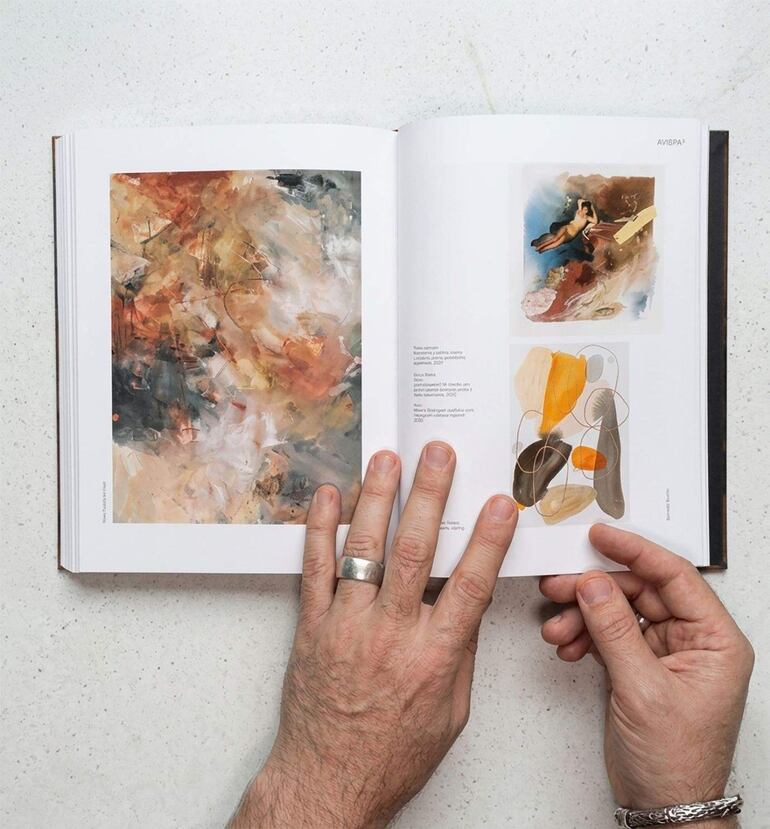 Manos sosteniendo un libro abierto que exhibe obras de arte abstractas en un ambiente de exposición.