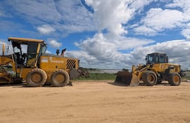 Máquina niveladora Volvo y retroexcavadora Komatsu en terreno rural con cielo despejado y vegetación al fondo.