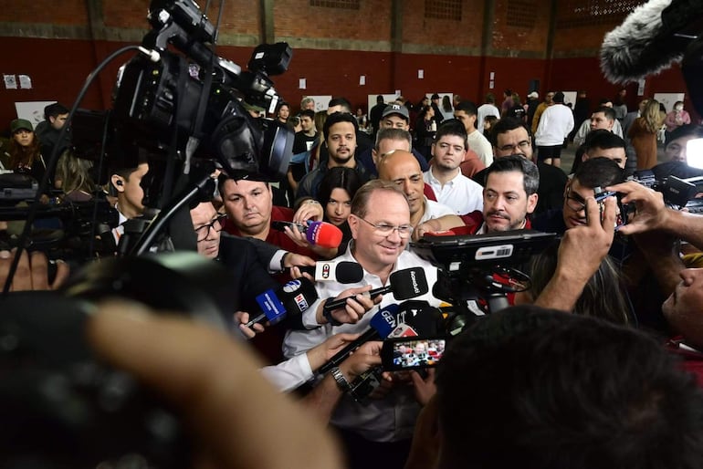 Camilo Pérez con camisa blanca habla a varios periodistas, quienes sostienen micrófonos y cámaras en un ambiente caótico.