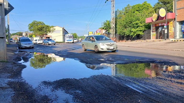 Calle con un bache profundo y agua acumulada, coches estacionados a los lados y vegetación en un día soleado.