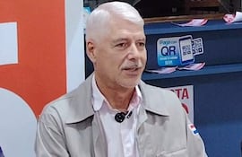 Arnaldo Wiens, con chaqueta clara, habla en un evento, mostrando un distintivo en la manga y en un ambiente con tecnología QR.