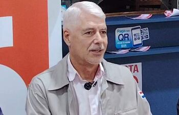 Arnaldo Wiens, con chaqueta clara, habla en un evento, mostrando un distintivo en la manga y en un ambiente con tecnología QR.