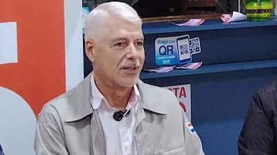 Arnaldo Wiens, con chaqueta clara, habla en un evento, mostrando un distintivo en la manga y en un ambiente con tecnología QR.