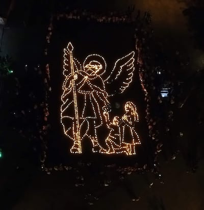 Instalación luminosa en forma de ángel que custodia a dos niños, rodeada de una multitud en un ambiente festivo y nocturno.