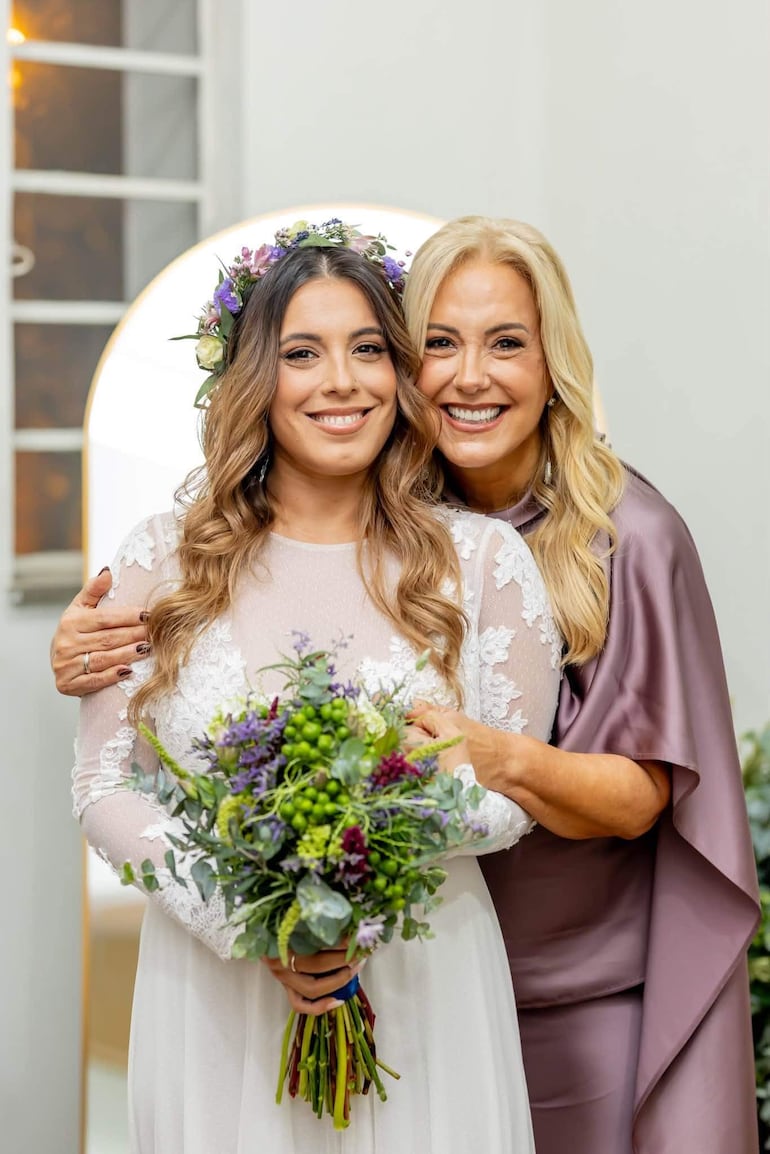 Menchi Barriocanal sonríe con vestido blanco y ramo de flores, mientras abraza a Mercedes Barriocanal, vestida de púrpura, en celebración.