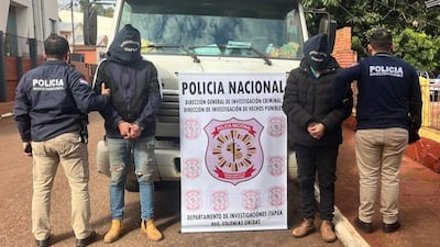 Dos hombres esposados con sudaderas, rodeados de policías con chaquetas identificativas, en una escena de detención policial.