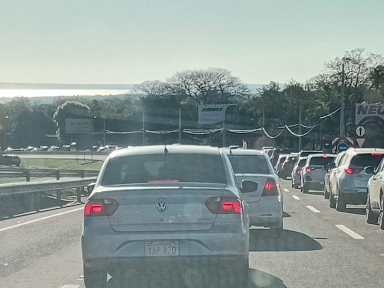 Varios vehículos en fila en un día soleado, sin personas fuera de los coches, paisaje con vegetación y un cuerpo de agua al fondo.