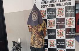 Joven de espaldas con camiseta camuflada y pañuelo oscuro en comisaría, rodeado de logotipos de la Policía Nacional.