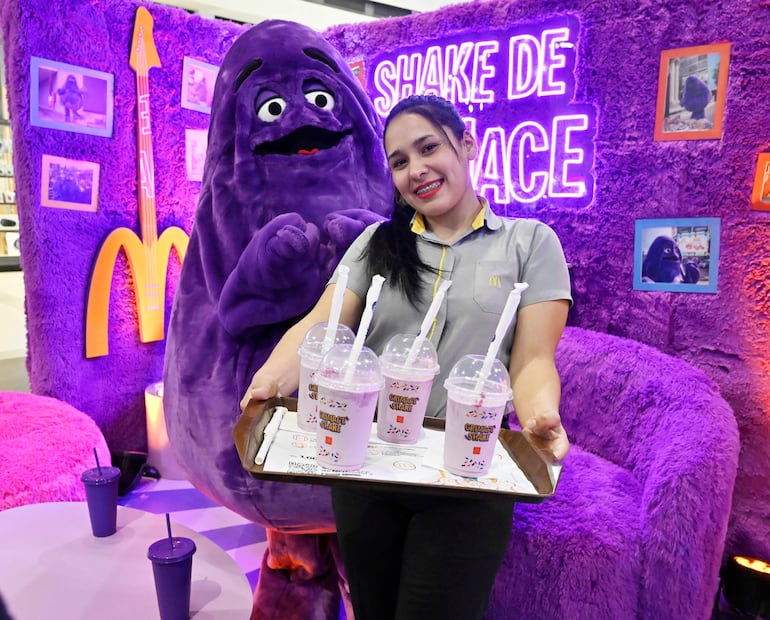 Mujer sonriente con camiseta de McDonald's sostiene bandeja de batidos, mientras Grimace posando detrás en ambiente decorado en morado.