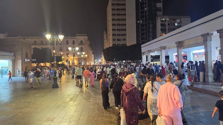 Multitud de personas de diversas etnias caminando y agrupándose en una plaza urbana iluminada claramente en la noche.