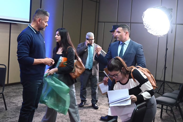 Hombre con suéter azul conversa con mujer de bolso marrón, mientras un hombre mayor usa su teléfono en un entorno iluminado.