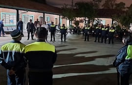 Grupo de policías en uniforme azul oscuro y amarillo, alineados en un espacio exterior iluminado con motocicletas estacionadas detrás.