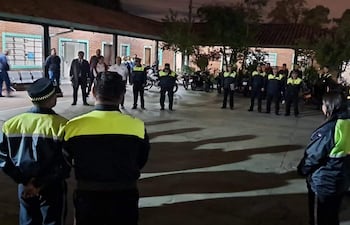 Grupo de policías en uniforme azul oscuro y amarillo, alineados en un espacio exterior iluminado con motocicletas estacionadas detrás.