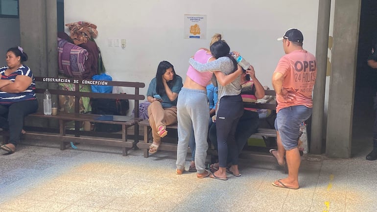 Mujeres abrazadas, una con top rosa y otra con suéter, en un ambiente sombrío de la Gobernación, observando la situación con tristeza.