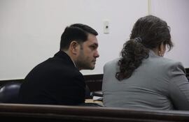 Hombre con chaqueta negra y mujer con chaqueta gris, ambos concentrados en documentos en tribunal.