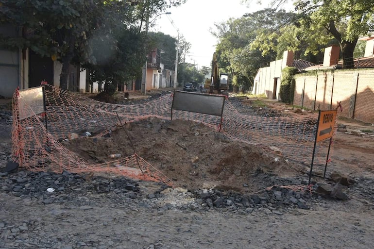 Agujero en carretera rodeado de malla de seguridad anaranjada, máquina excavadora al fondo y cartel de desvío visible.