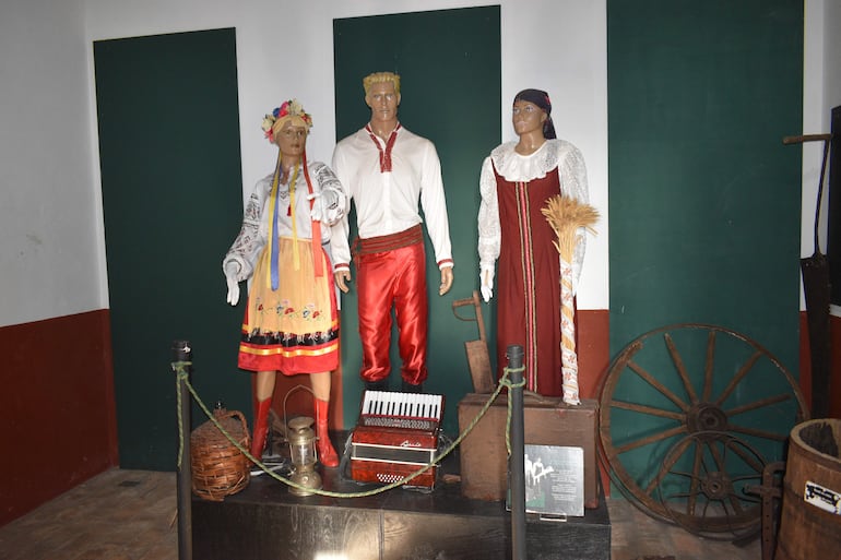 Tres maniquíes en museo, vestidos con trajes tradicionales, rodeados de objetos culturales como un acordeón y un carro de madera.