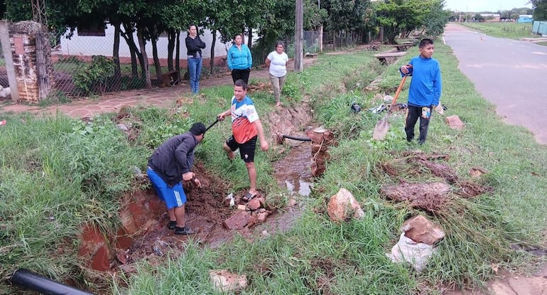 Dos hombres excavan un canal de drenaje, mientras un niño observa y tres mujeres miran con interés. Vegetación en el fondo.